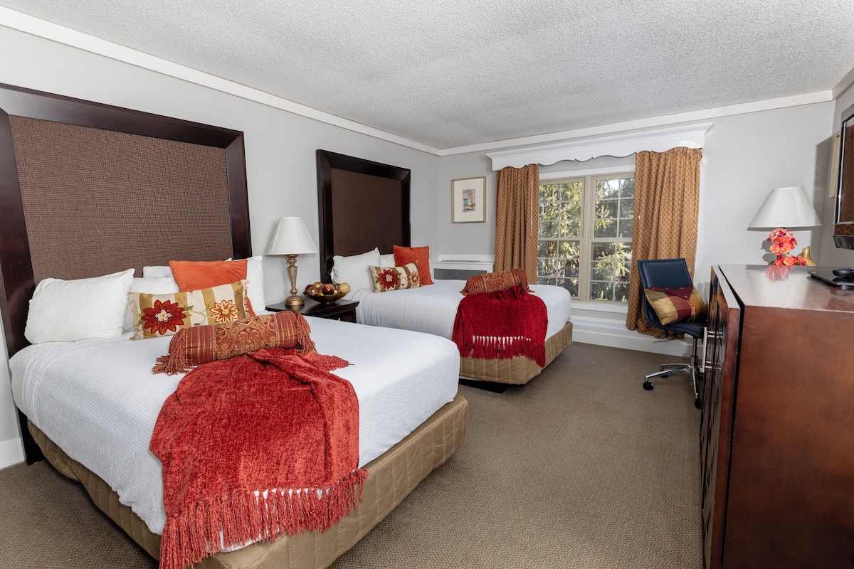 A hotel room with two beds, a desk, a chair, a lamp, and a window with curtains.