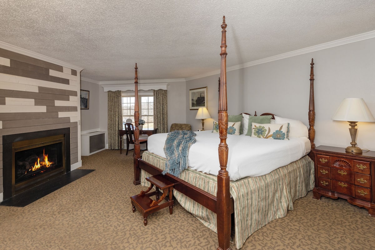 A four-poster bed with a wooden frame, a fireplace, a bedside table, and a nightstand.