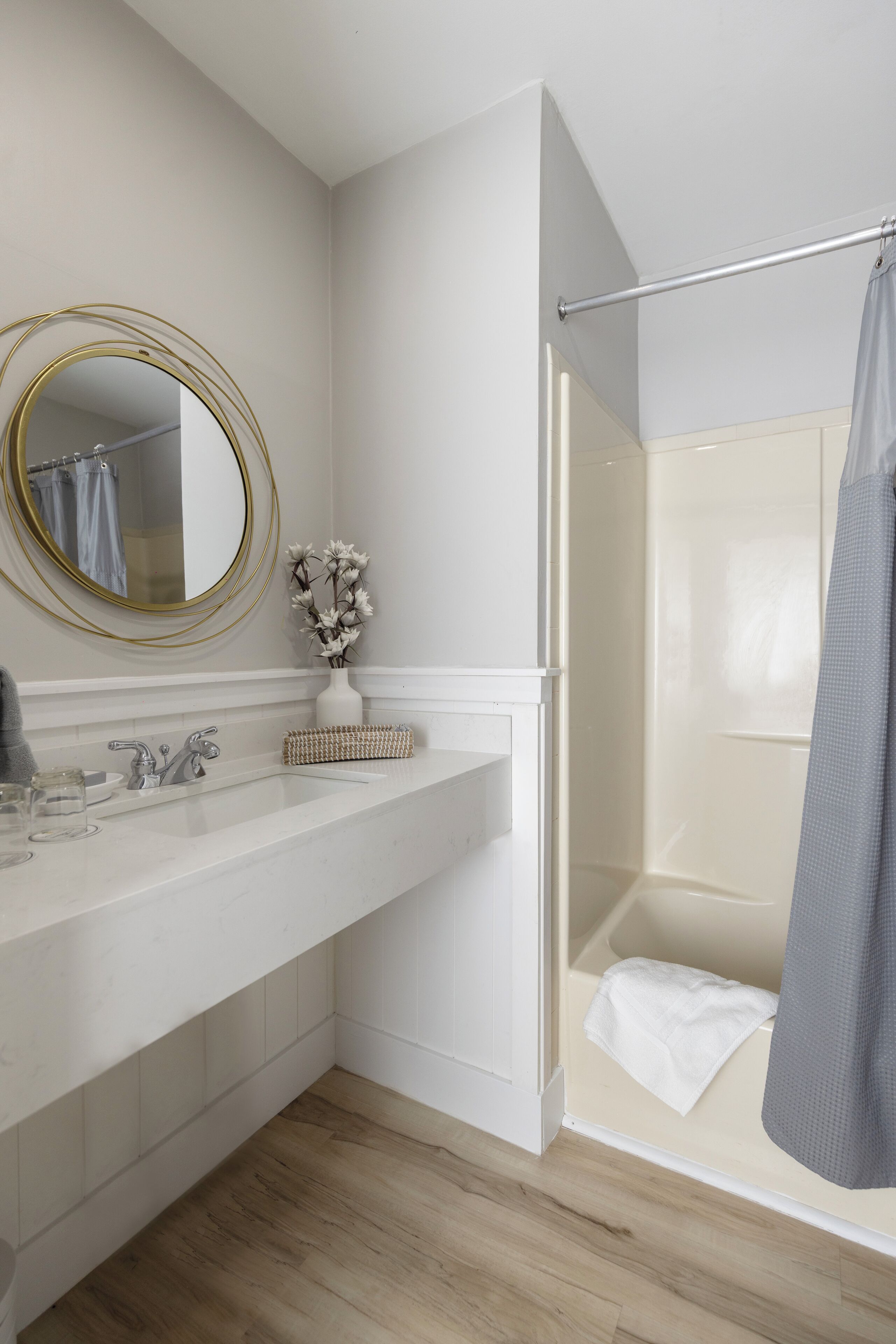 A bathroom with a white vanity, a gold-framed mirror, a vase with flowers, and a shower area with a towel.