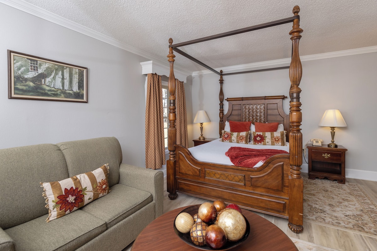 A four-poster bed with a wooden headboard, a sofa with a floral pillow, a bedside table with a lamp, and a painting on the wall.