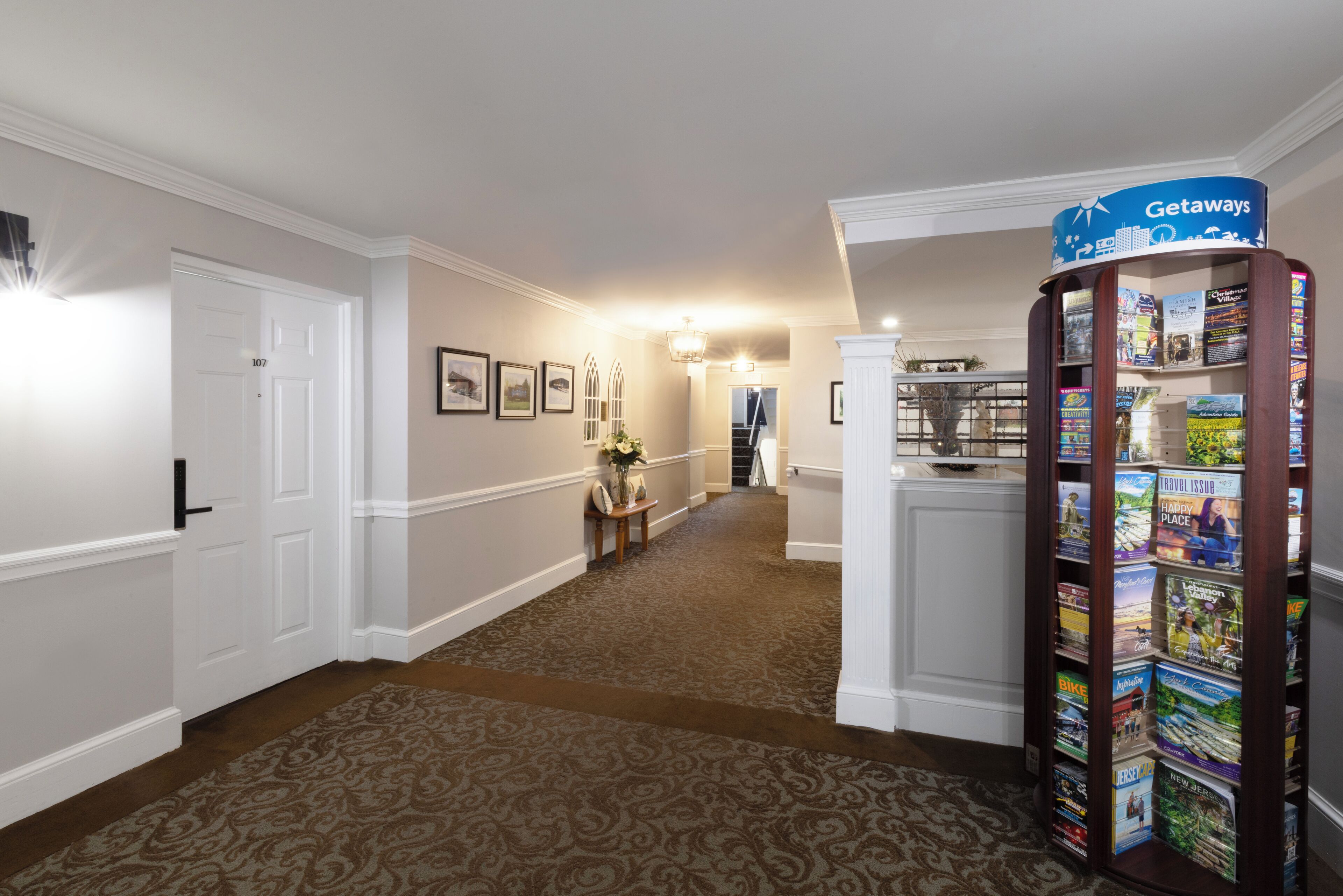 A hallway with a patterned carpet, a reception desk, a 'Getaways' magazine rack, and framed pictures on the wall.