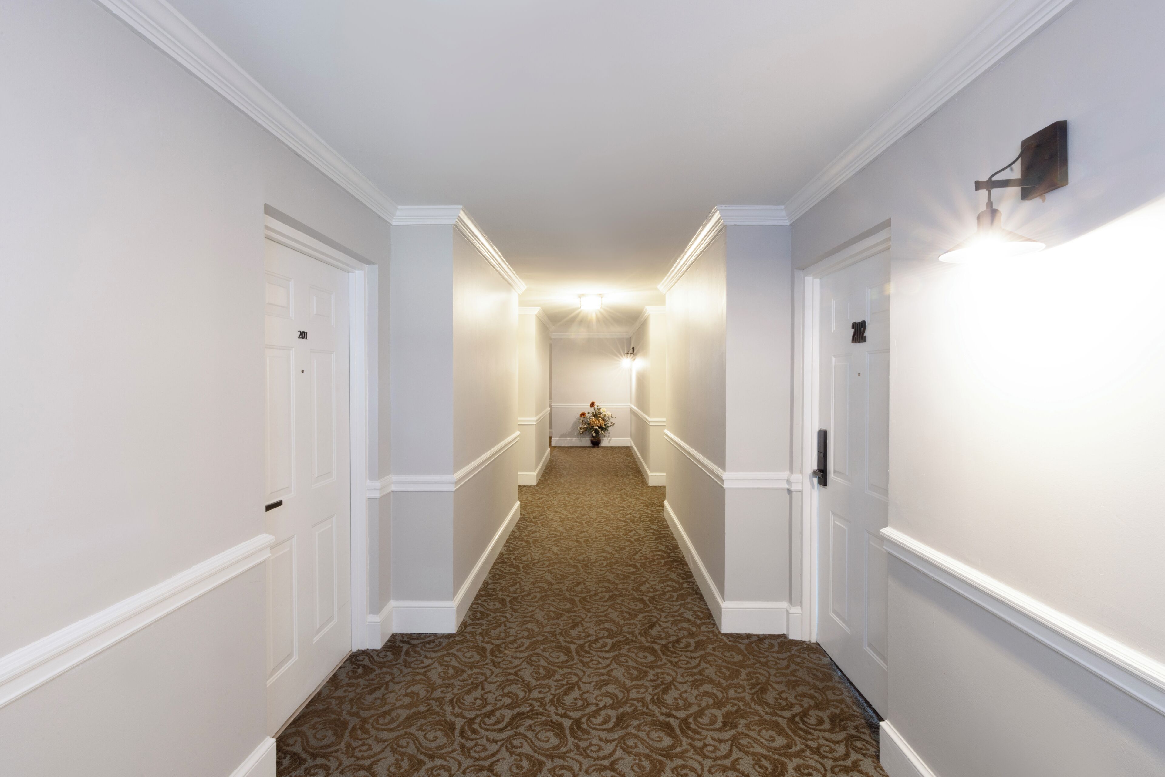 A hallway with a patterned carpet, white walls, and a light fixture at the end.