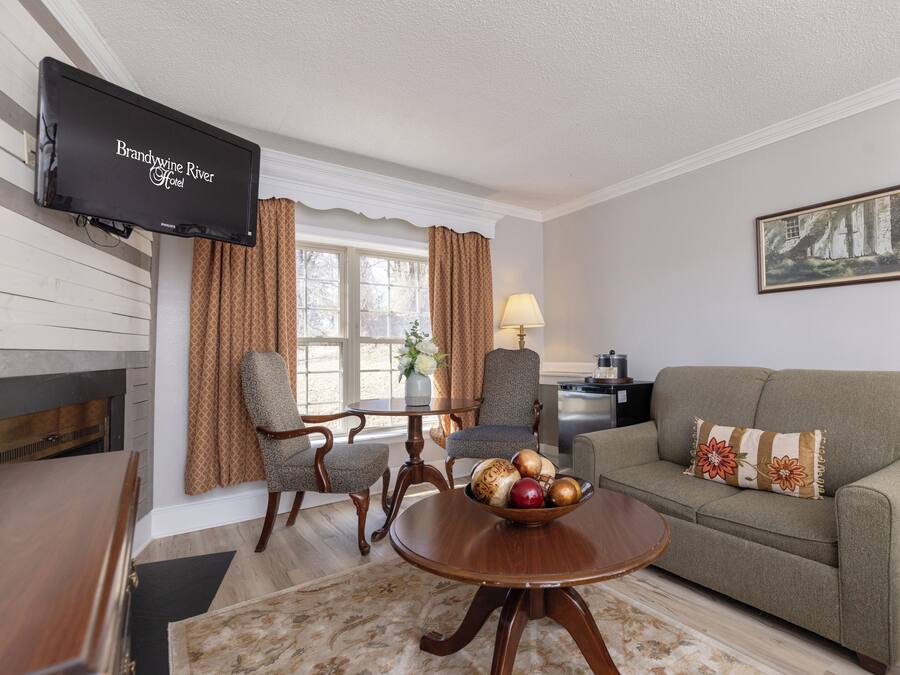 A living room with a television displaying 'Brandywine River Hotel', a sofa, armchairs, a wooden coffee table with fruit, and a painting on the wall.