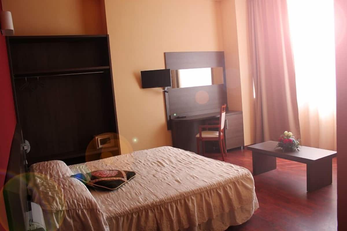 A hotel room with a bed, a desk, a chair, a TV, and a small table with a flower arrangement.