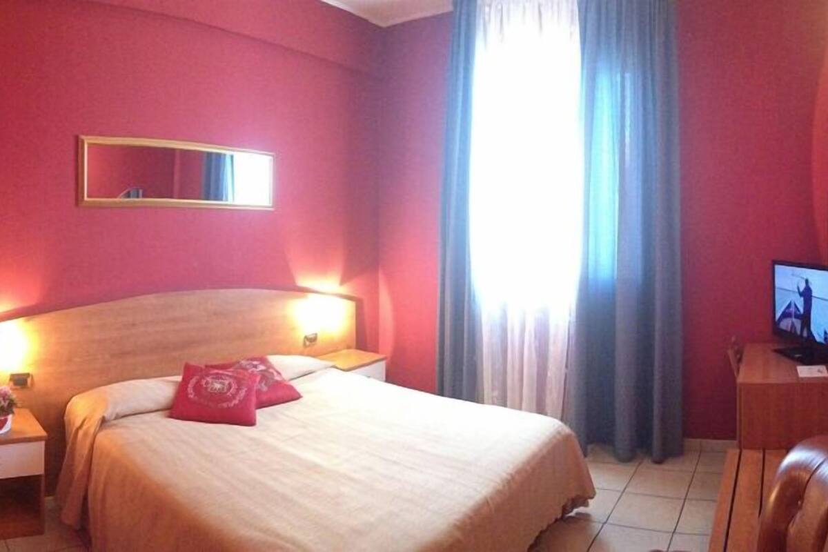 A hotel room with a bed, bedside lamps, a TV, and a window with curtains.