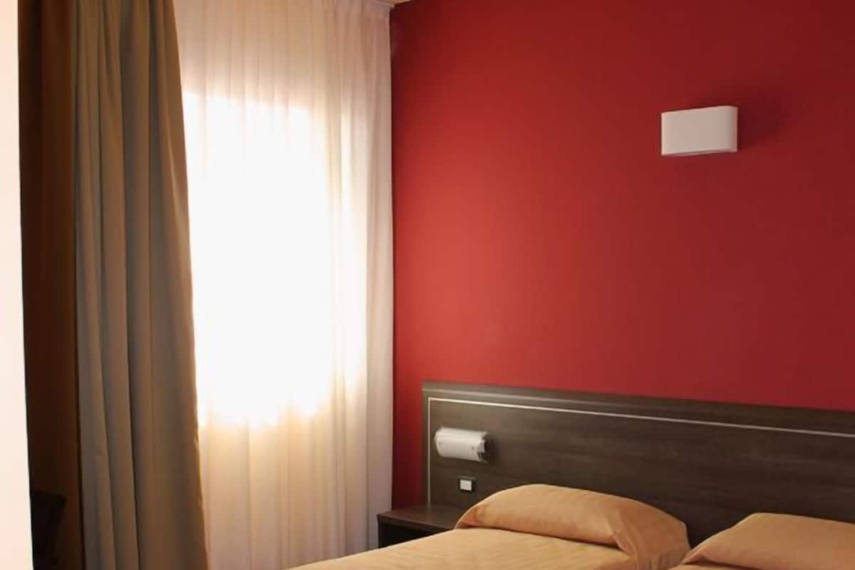 A room with two single beds, a wooden headboard, a red wall, and a window with curtains.