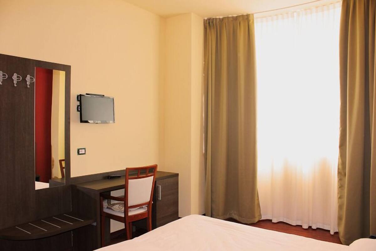 A hotel room with a bed, a desk with a chair, a television, and a window with curtains.