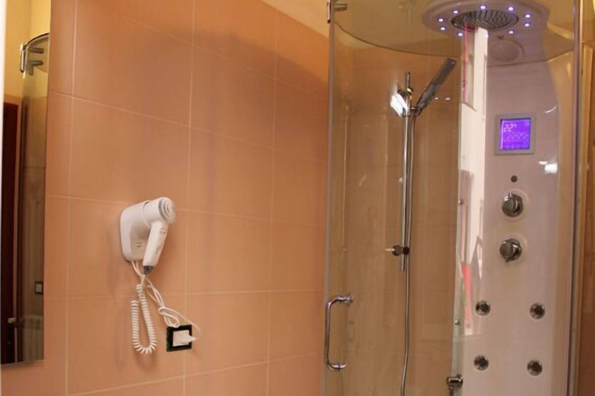A glass-enclosed shower stall with a handheld showerhead, a control panel, and a small step stool.