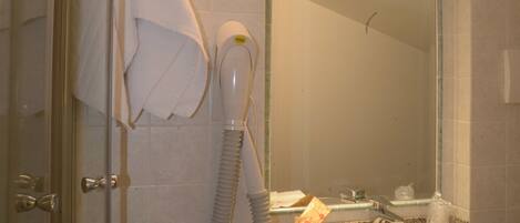 A bathroom with a glass shower door, a white towel hanging on the door, a hairdryer, and a sink with a mirror above it.