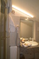Un baño con mampara de vidrio, una toalla blanca colgada en la puerta, un secador de pelo y un lavabo con un espejo encima.