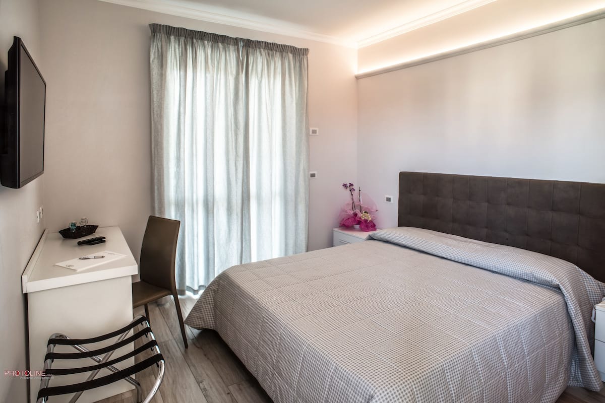 A hotel room with a bed, a desk with a chair, a TV mounted on the wall, and a window with curtains.