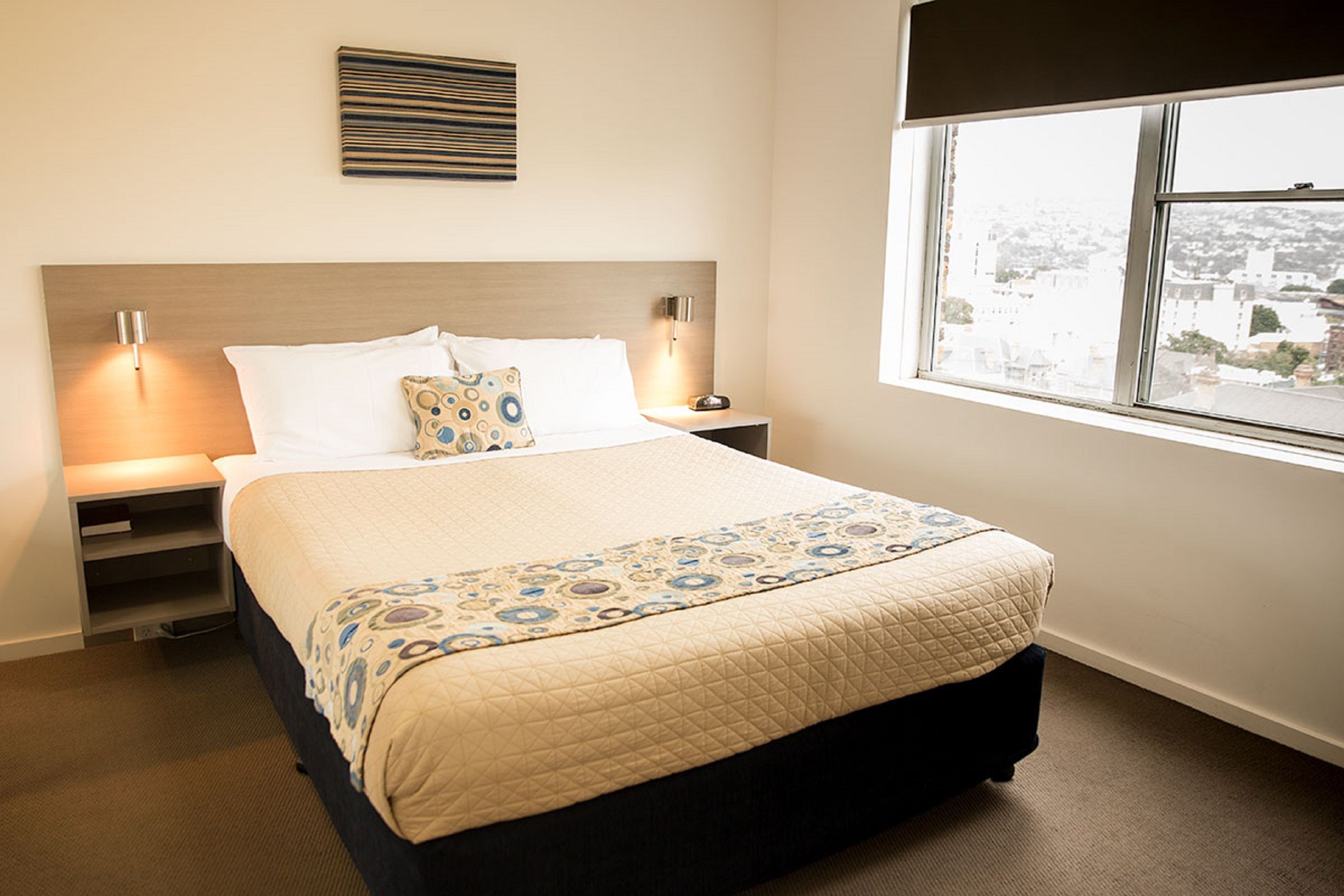Una cama bien tendida con una colcha estampada, una lámpara de noche y una ventana con vista a la ciudad.
