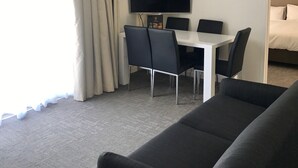 A hotel room with a large window, a flat-screen TV mounted on the wall, a dining table with chairs, a sofa, and a bed with a headboard. - Adina Place Motel Apartments (Launceston)
