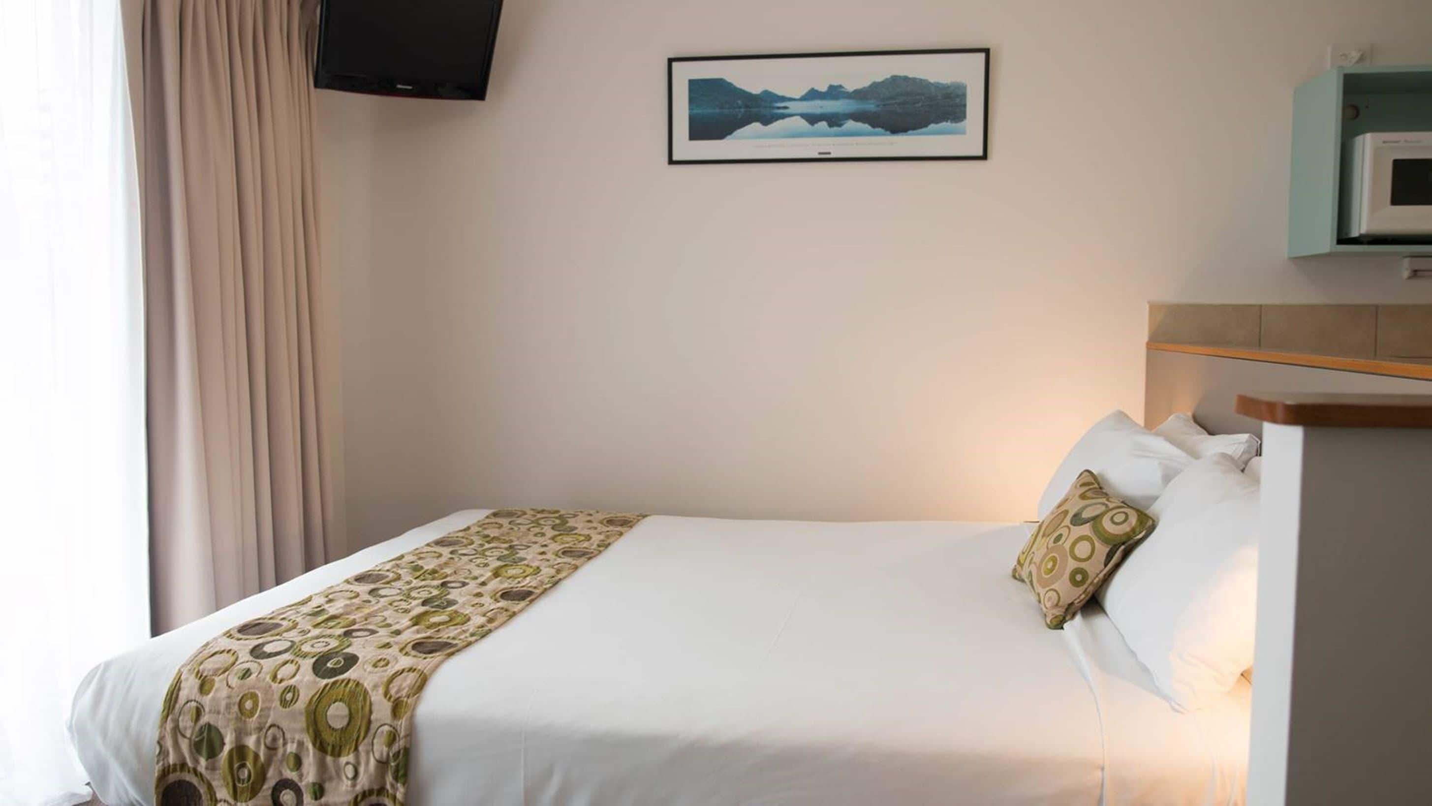 A hotel room with a bed, a mounted TV, a framed picture on the wall, and a small shelf with a microwave.
