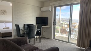 A hotel room with a bed, a desk with a chair, a television, and a balcony with a view of the city. - Adina Place Motel Apartments (Launceston)
