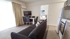 A modern living room with a dark sofa, a dining table with chairs, a flat-screen TV, and a kitchen area with a refrigerator and oven. - Adina Place Motel Apartments (Launceston)