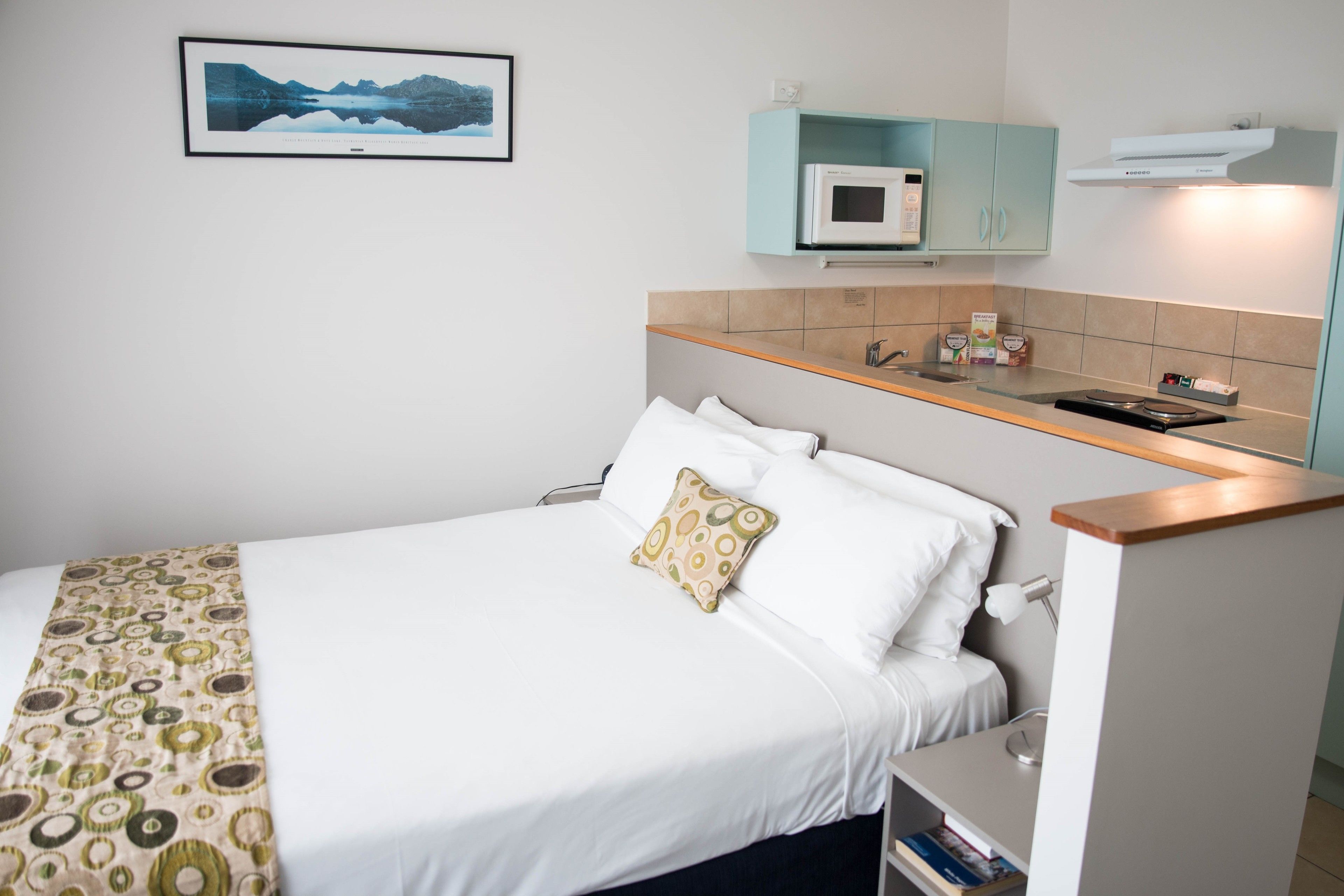 A neatly made bed with a patterned blanket, a bedside table with a lamp, and a microwave above a light blue cabinet.