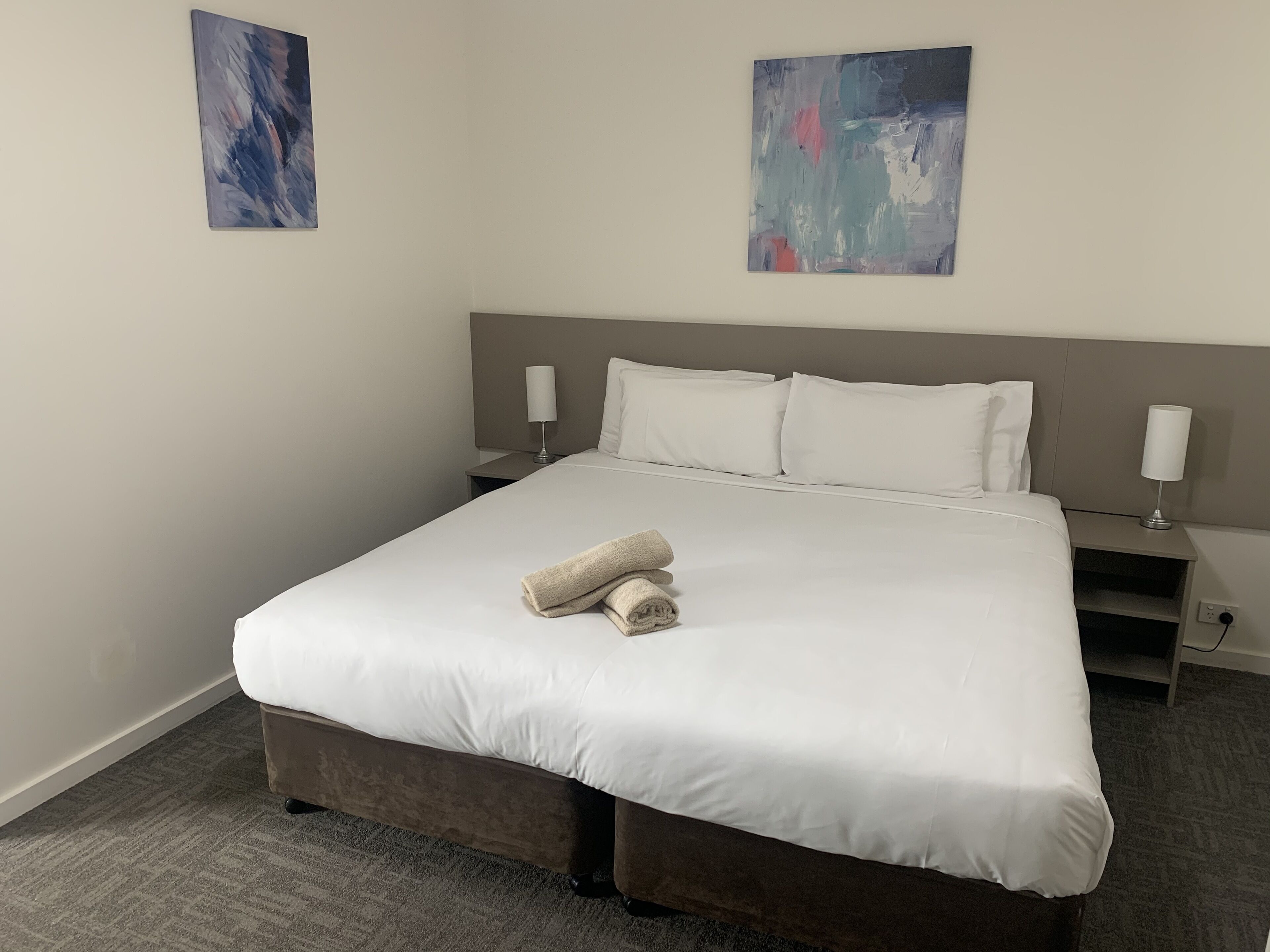 A neatly made bed with white linens, two bedside tables with lamps, and abstract art on the walls.