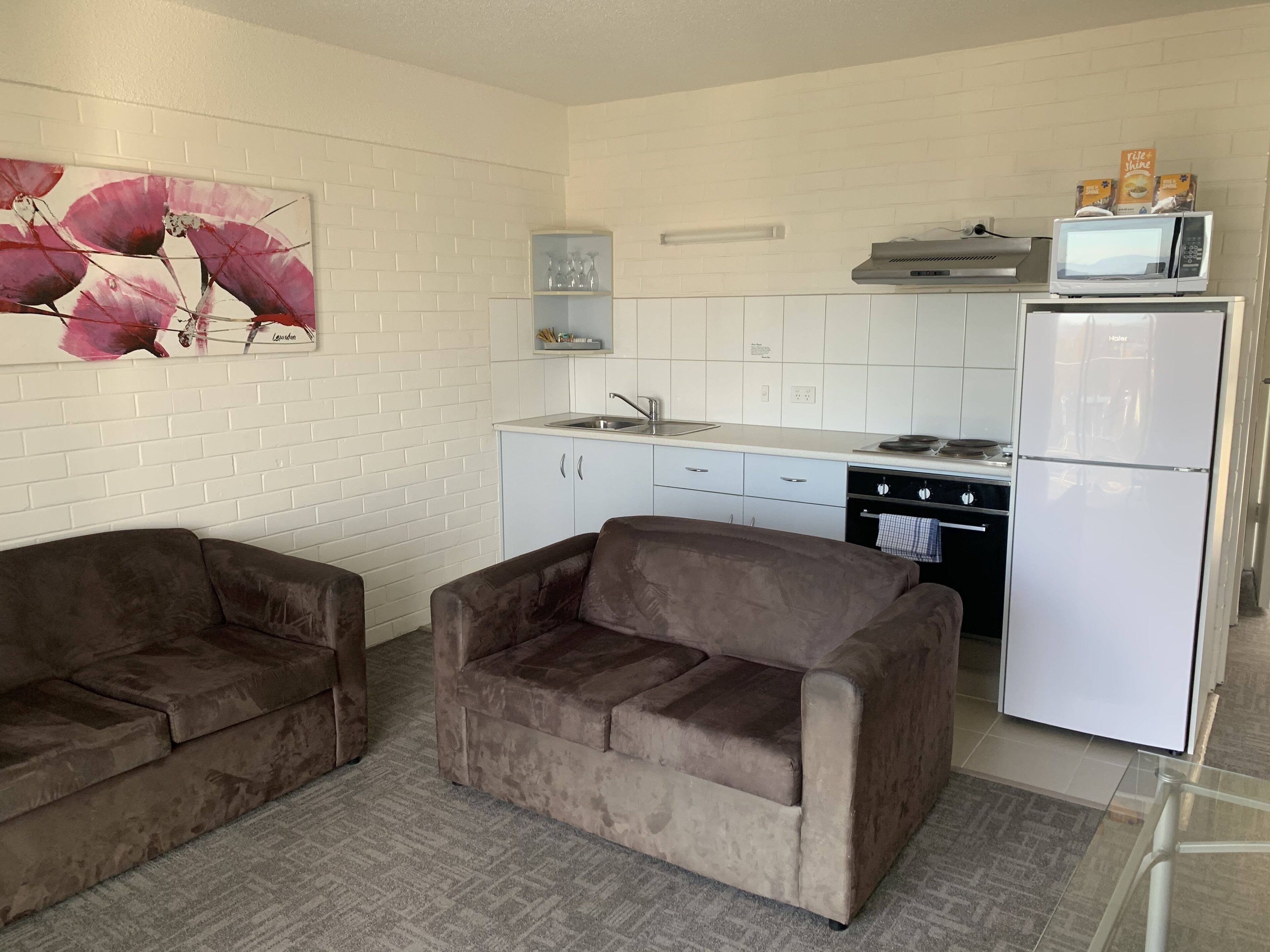 A compact kitchen with white brick walls, a microwave, a refrigerator, and a dishwasher. There are two brown sofas and a painting of pink flowers on the wall.