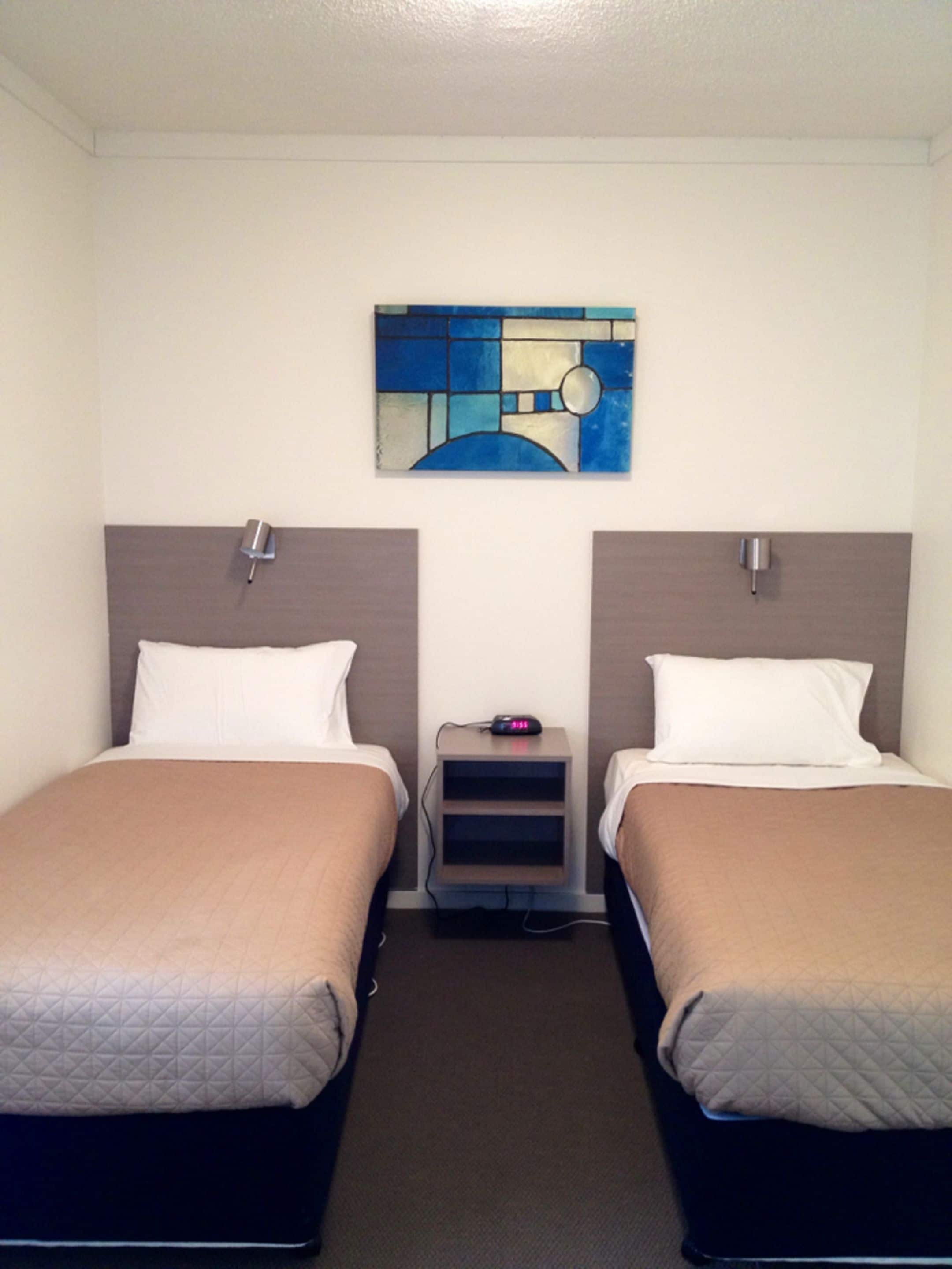 A hotel room with two beds, a small bedside table with a clock, and a wall art piece above the beds.