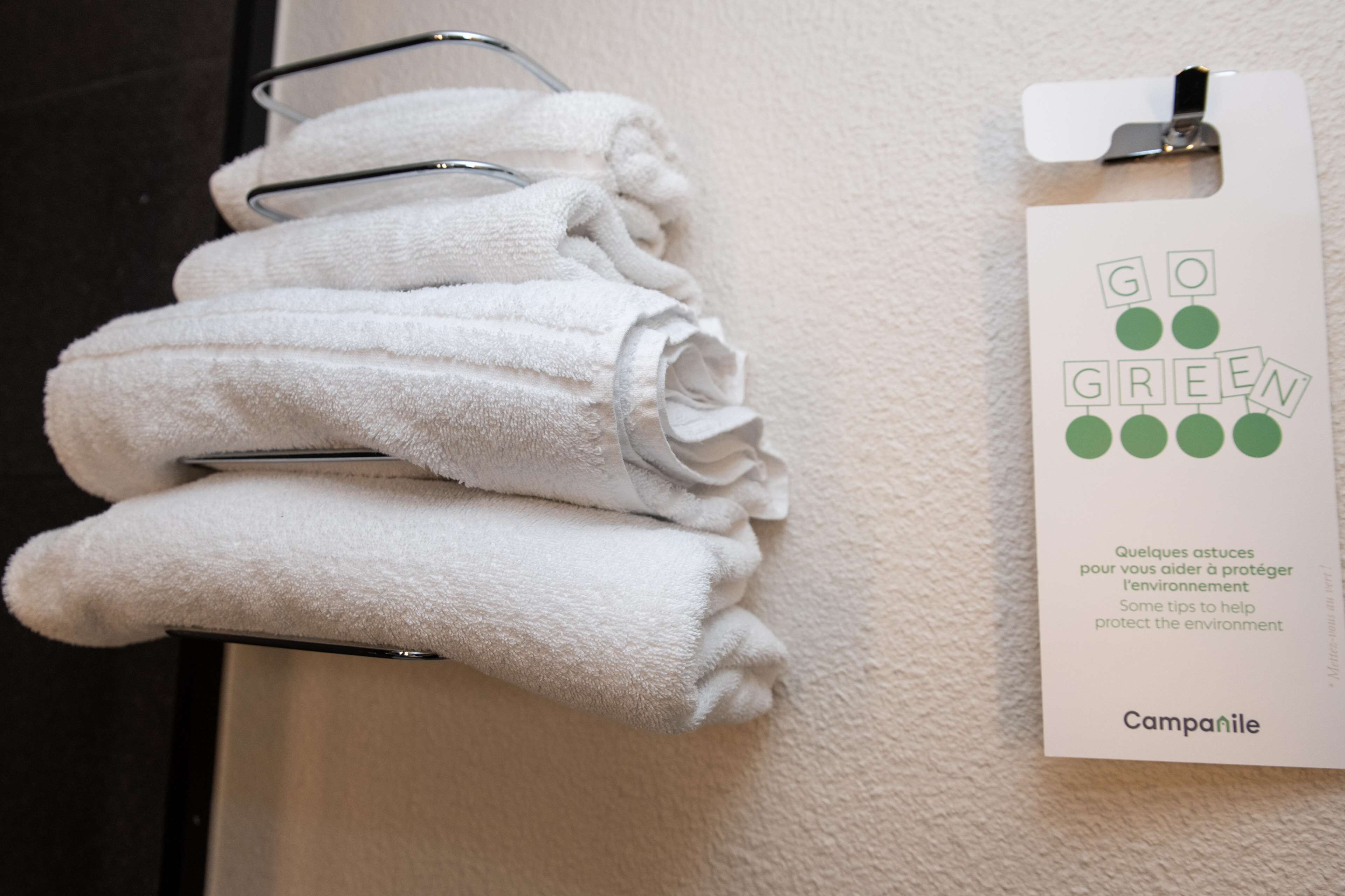 Una pila de toallas blancas sobre un esquinero metálico, junto a una pared, con una etiqueta blanca que dice “GO GREEN” e incluye consejos para el cuidado del medio ambiente.