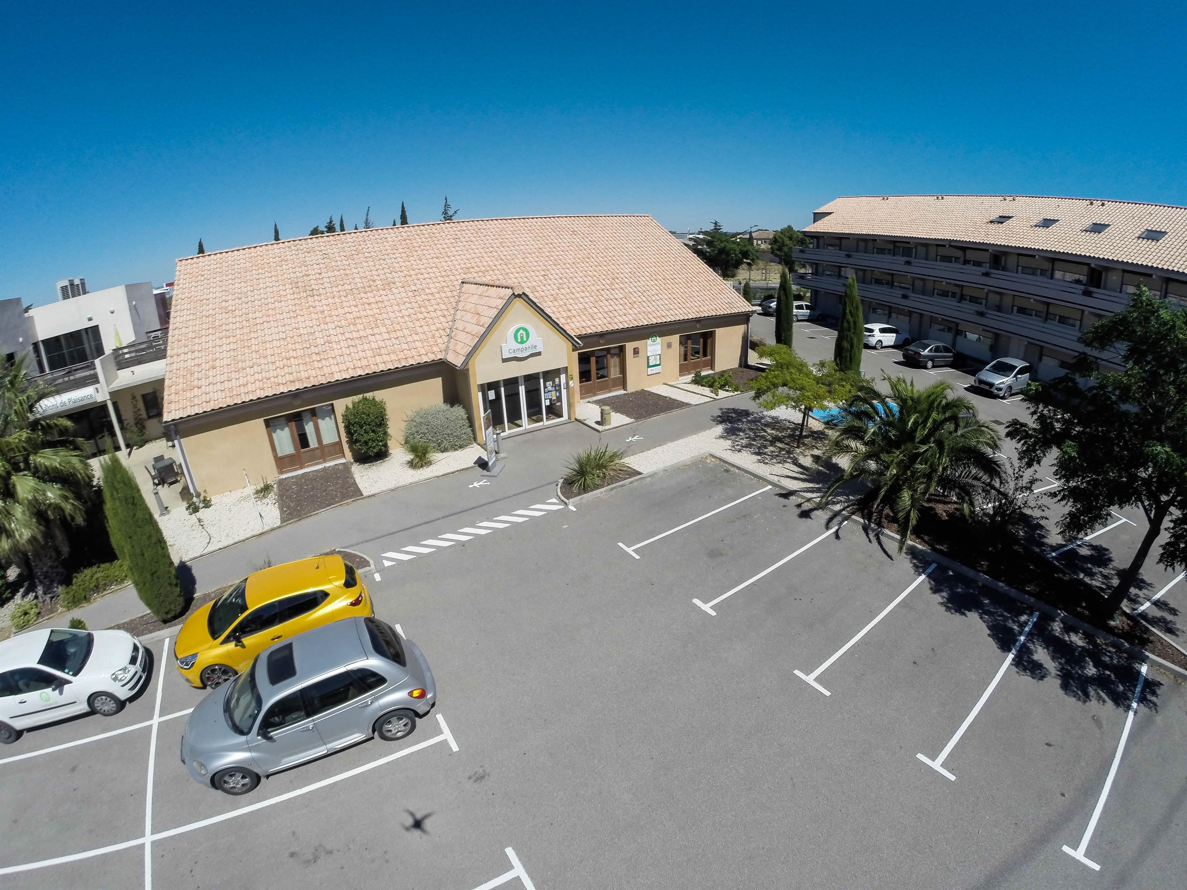 Un hôtel avec un toit recouvert de tuiles, un parking avec des places délimitées et un bâtiment sur lequel on peut lire l’inscription « Campanile ».