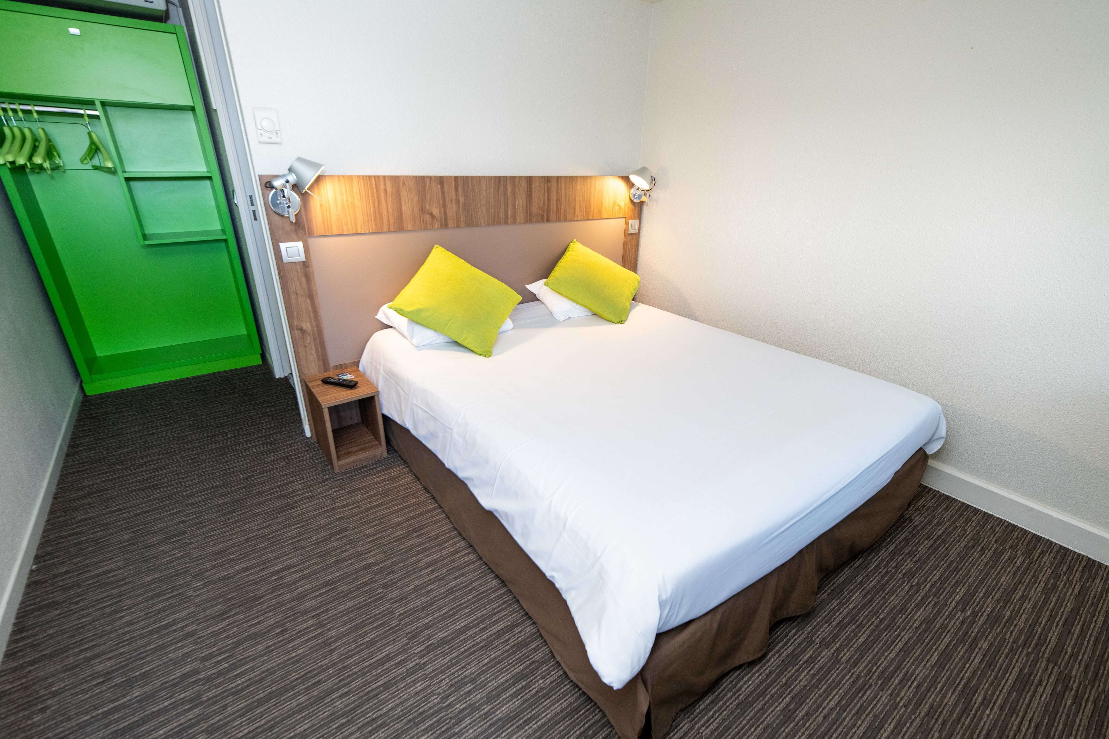 Une chambre d’hôtel avec un lit, une table de chevet, une armoire verte et une lampe fixée au mur.