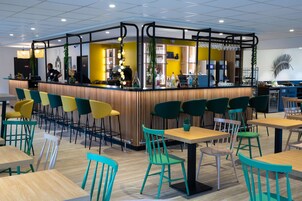 Un café moderno con mostrador de madera, sillas verdes y amarillas, y una zona de bar.