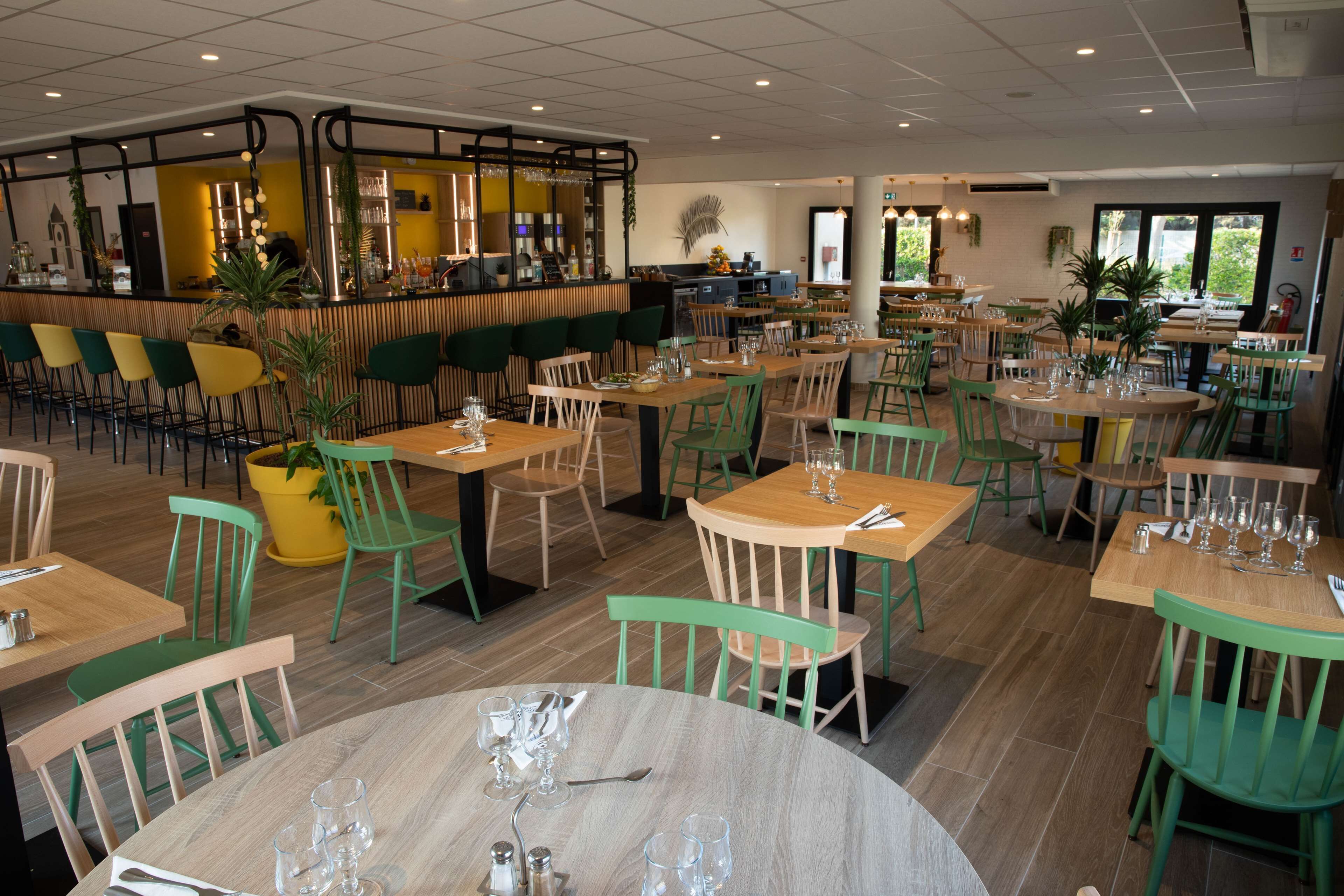 Een ruim restaurant met meerdere tafels en stoelen, een bar en potplanten.