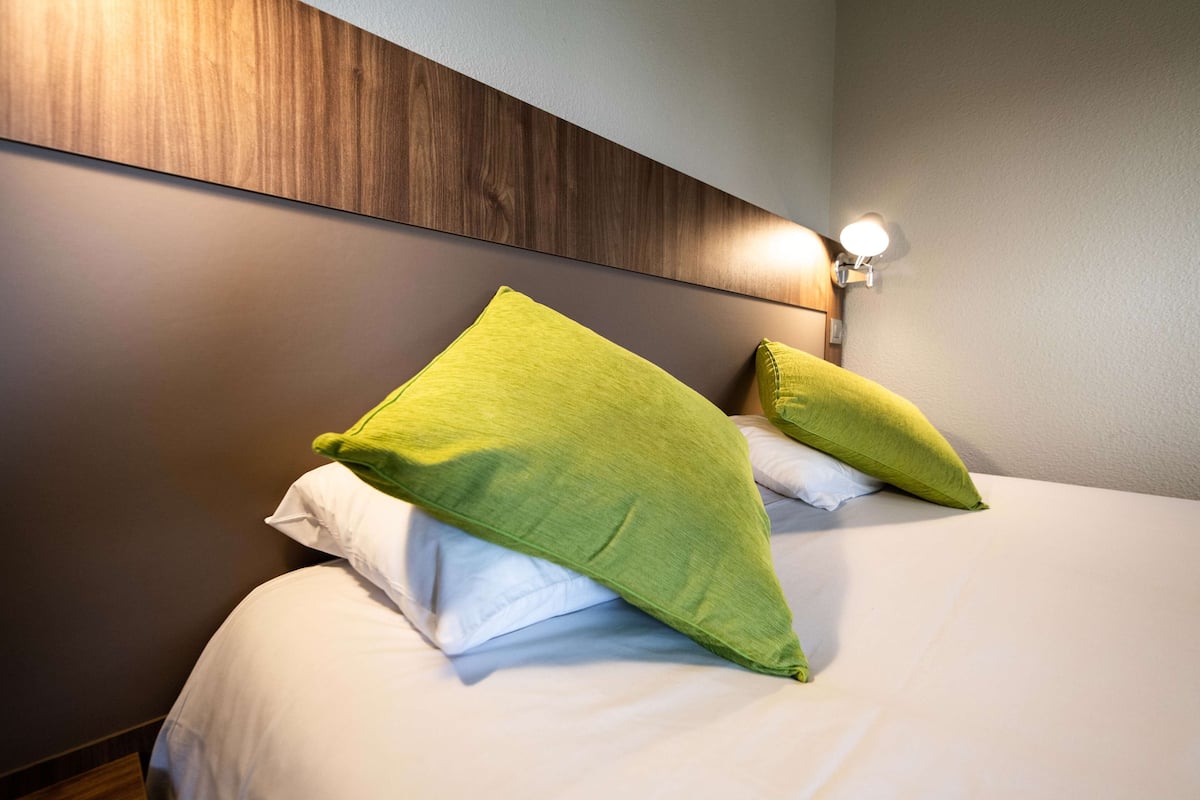 Una cama bien hecha con dos almohadas y una lámpara fijada a la pared.
