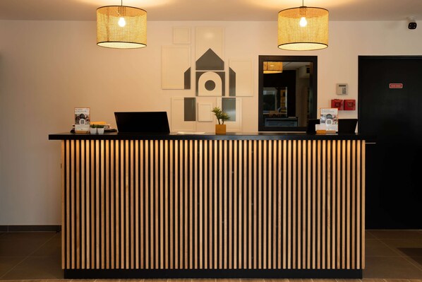 A reception desk with vertical wooden slats, two hanging wicker lamps, and framed artwork on the wall. - Campanile Narbonne (Narbonne)