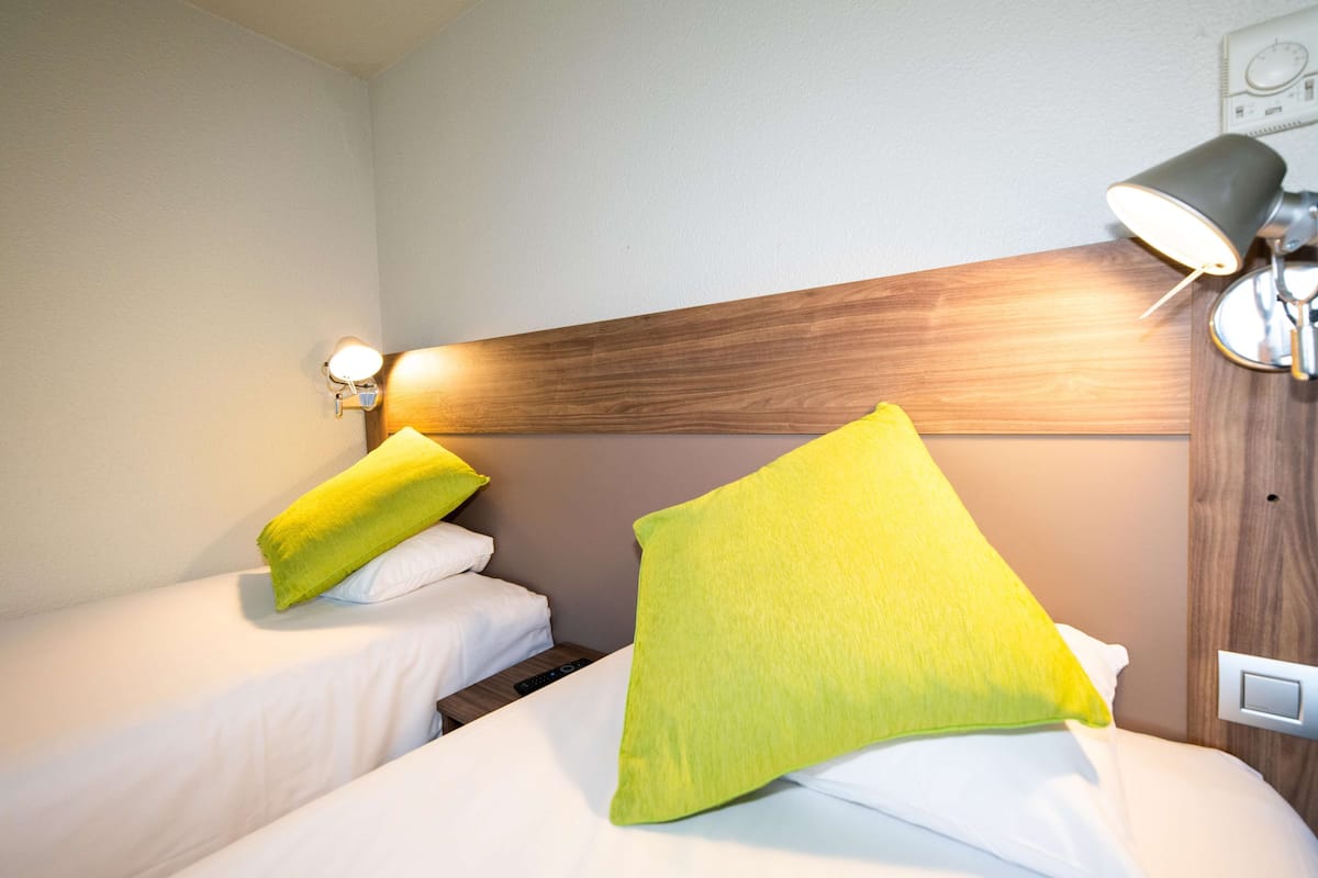 Habitación de hotel con dos camas individuales, cada una con un colchón blanco y un almohadarilla amarillo brillante. Tiene un cabecero de madera, una lámpara de pared y un enchufe en la pared.
