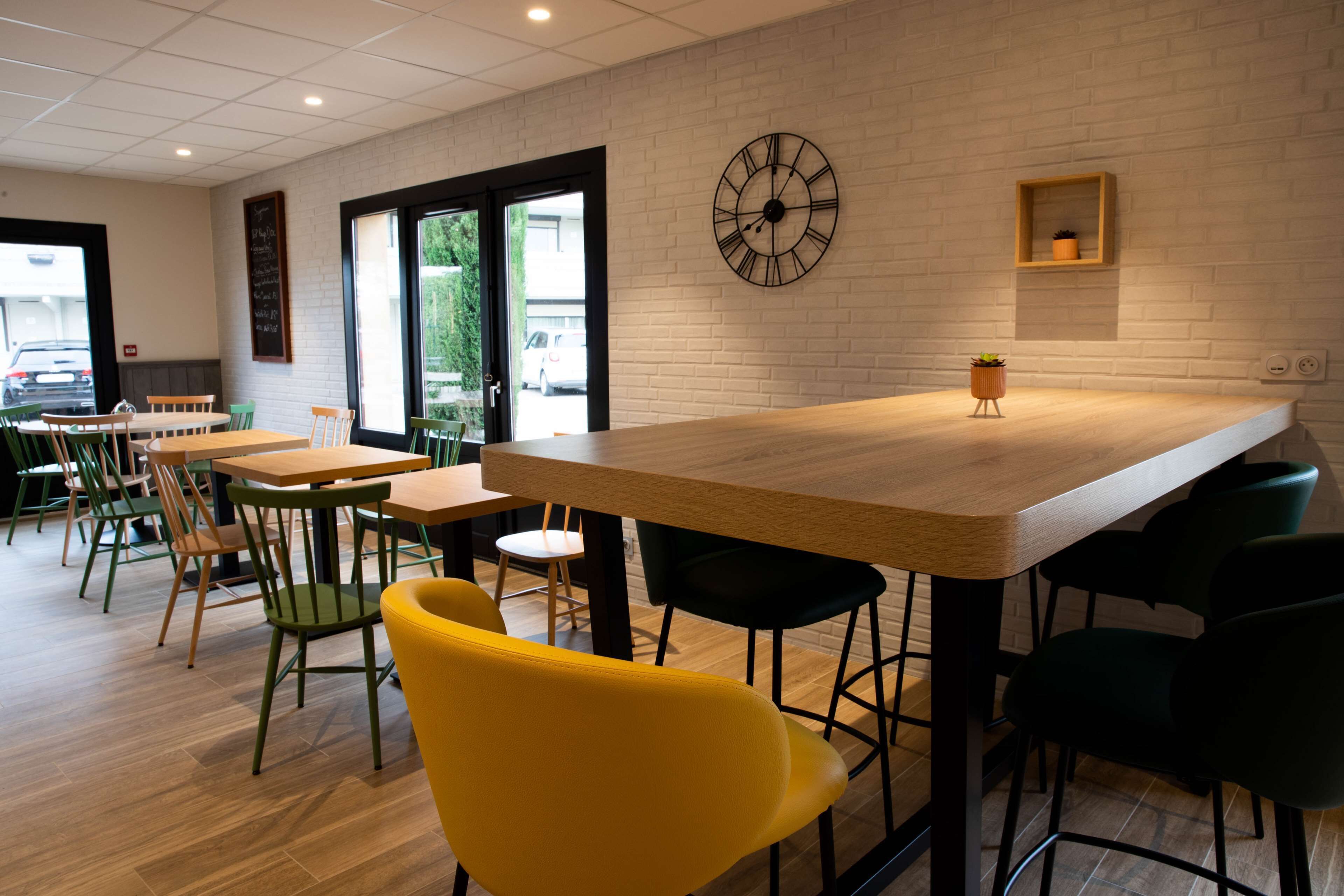 Ein modernes Café-Interieur mit Holztischen und -stühlen, einer großen Wanduhr und einem kleinen Regal mit einer Pflanze.