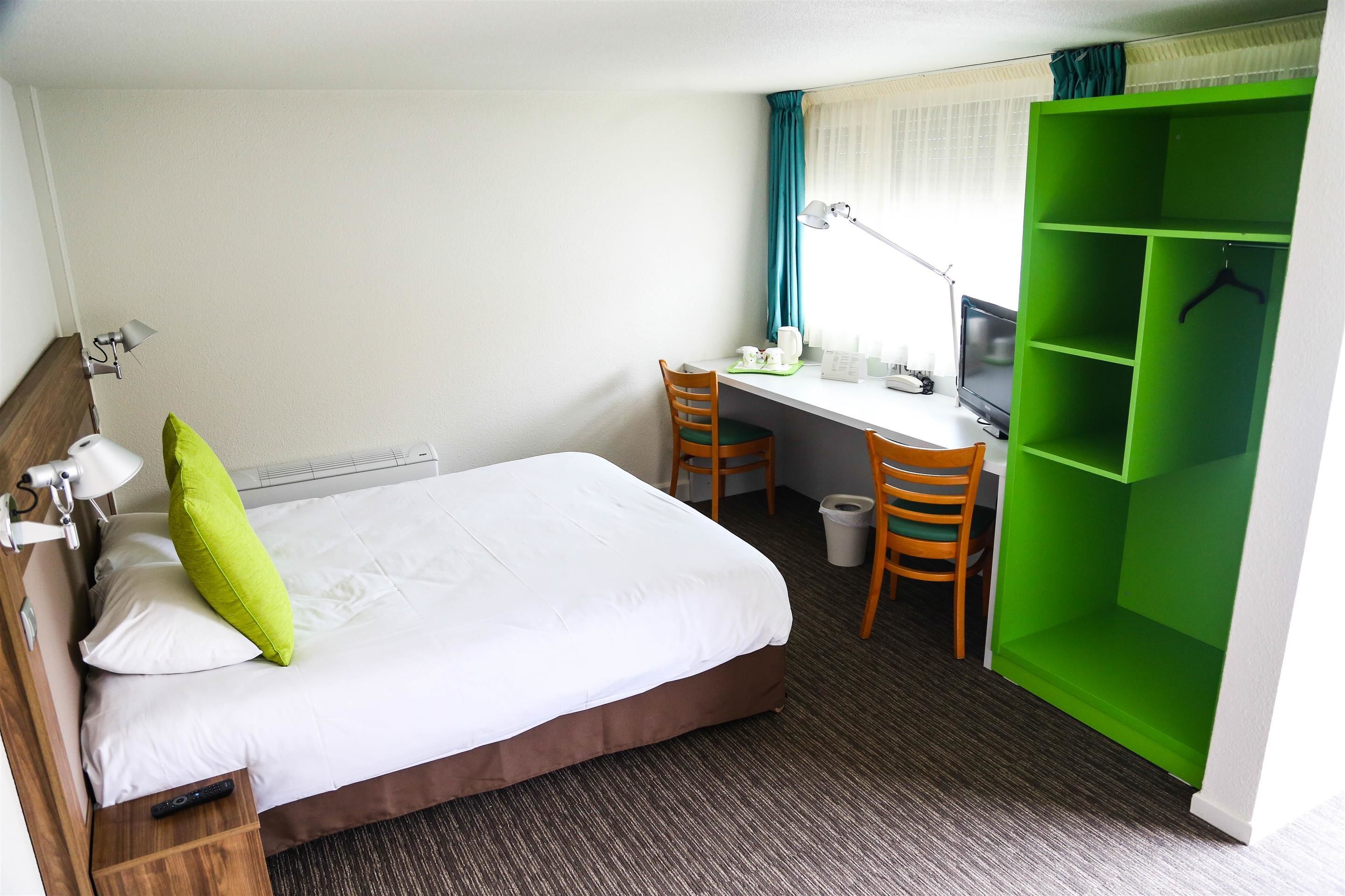 Habitación de hotel con cama, escritorio, sillas, armario verde y ventana con cortinas.