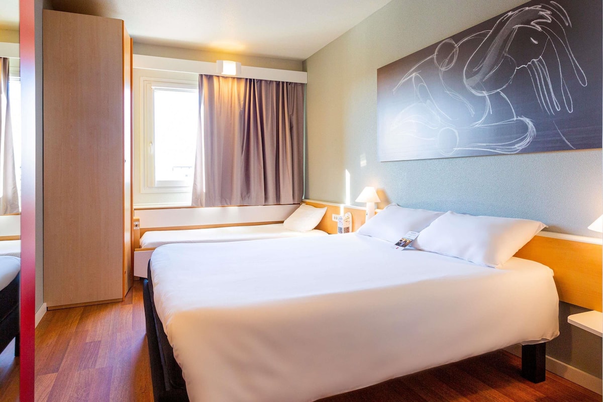 A hotel room with a large bed, bedside tables, a window with curtains, and a wall-mounted artwork.
