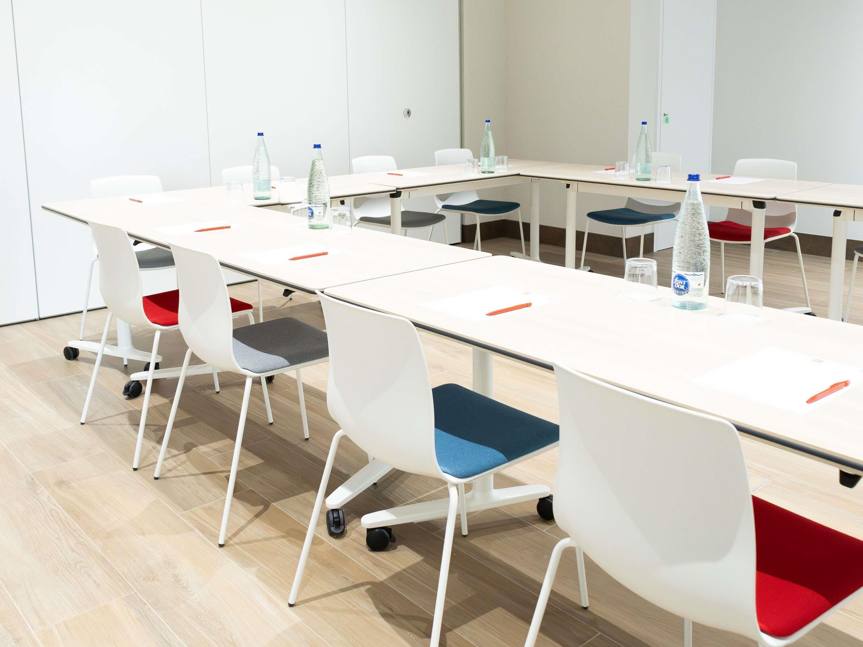 Een vergaderzaal met een lange tafel, witte stoelen met rode en blauwe kussens, en waterflesjes op de tafel.