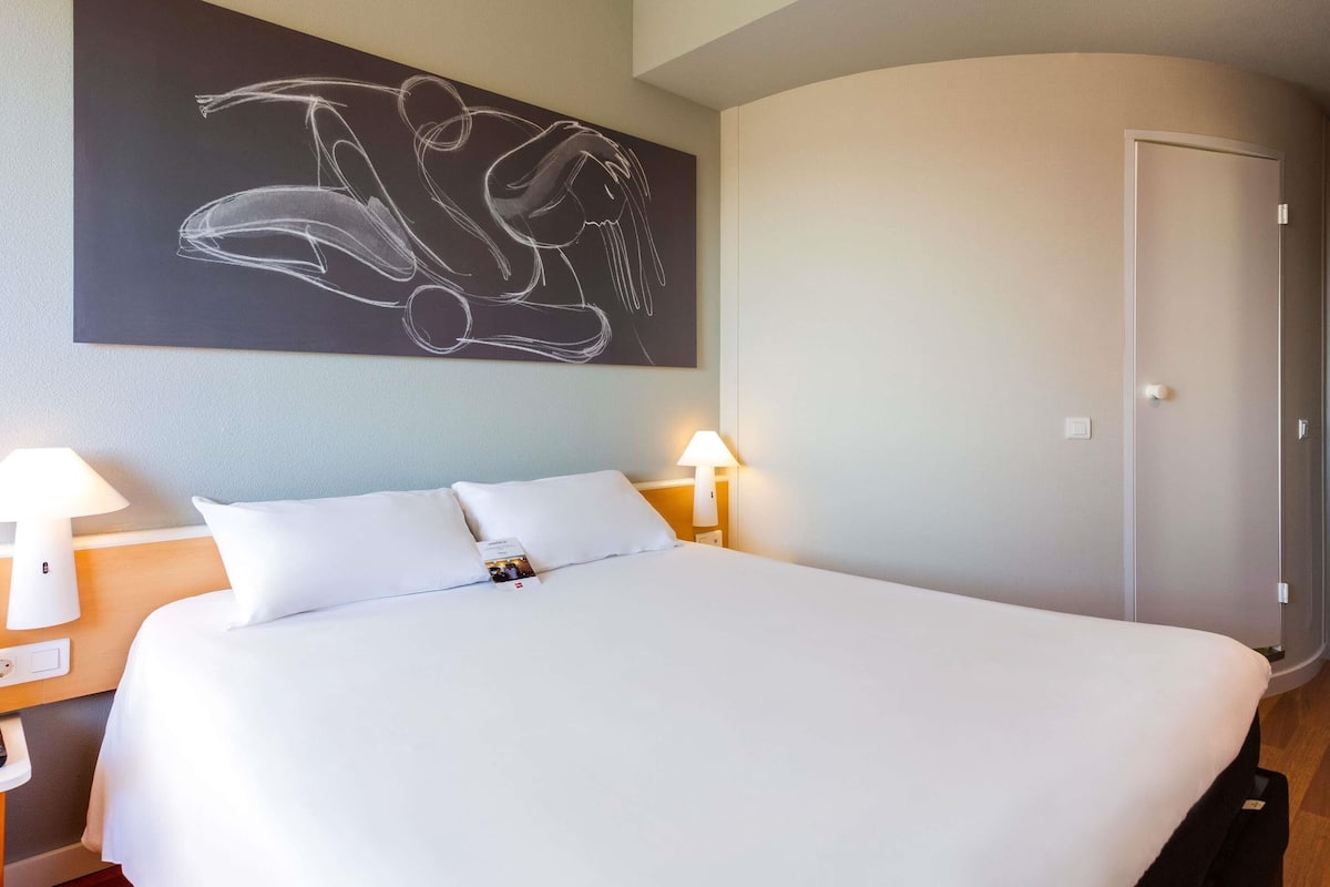 A hotel room with a large bed, two bedside lamps, a wall-mounted abstract artwork, and a door.