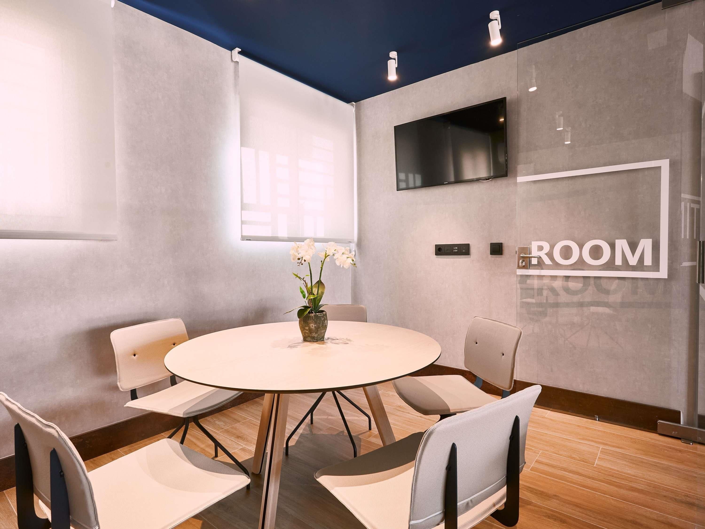 Een vergaderzaal met een ronde tafel, zes stoelen, een vaas met bloemen, een flatscreen-tv en een bordje met de tekst 'ROOM' aan de muur.