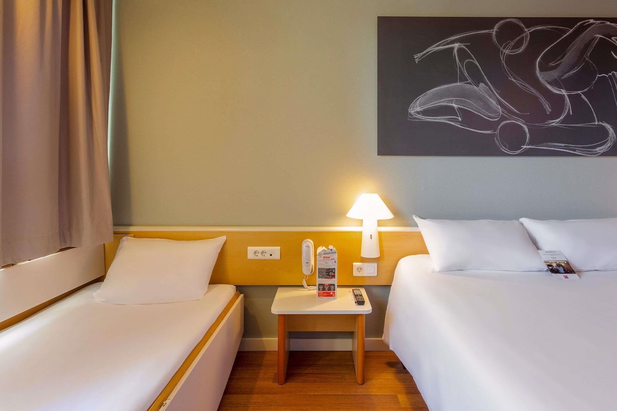 Hotellihuone, jossa on kaksi sänkyä, yöpöytä, lamppu, taulu ja näkymä ulos.