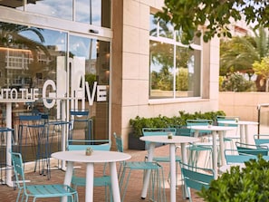 An outdoor seating area with tables and chairs, a building entrance with a glass door, and greenery.