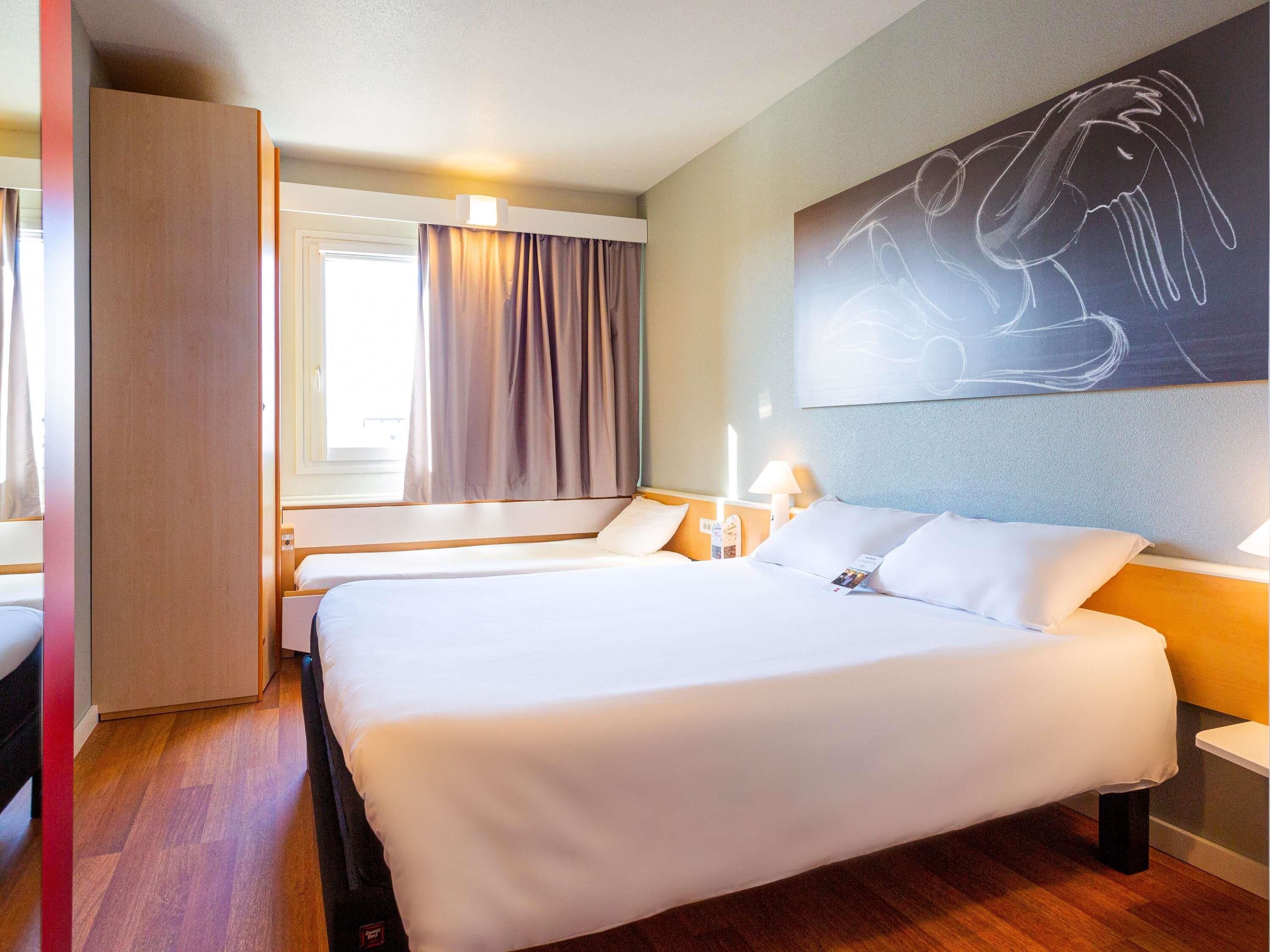 Habitación de hotel con una cama grande, mesitas de noche, una ventana con cortinas y una obra de arte colgada en la pared.