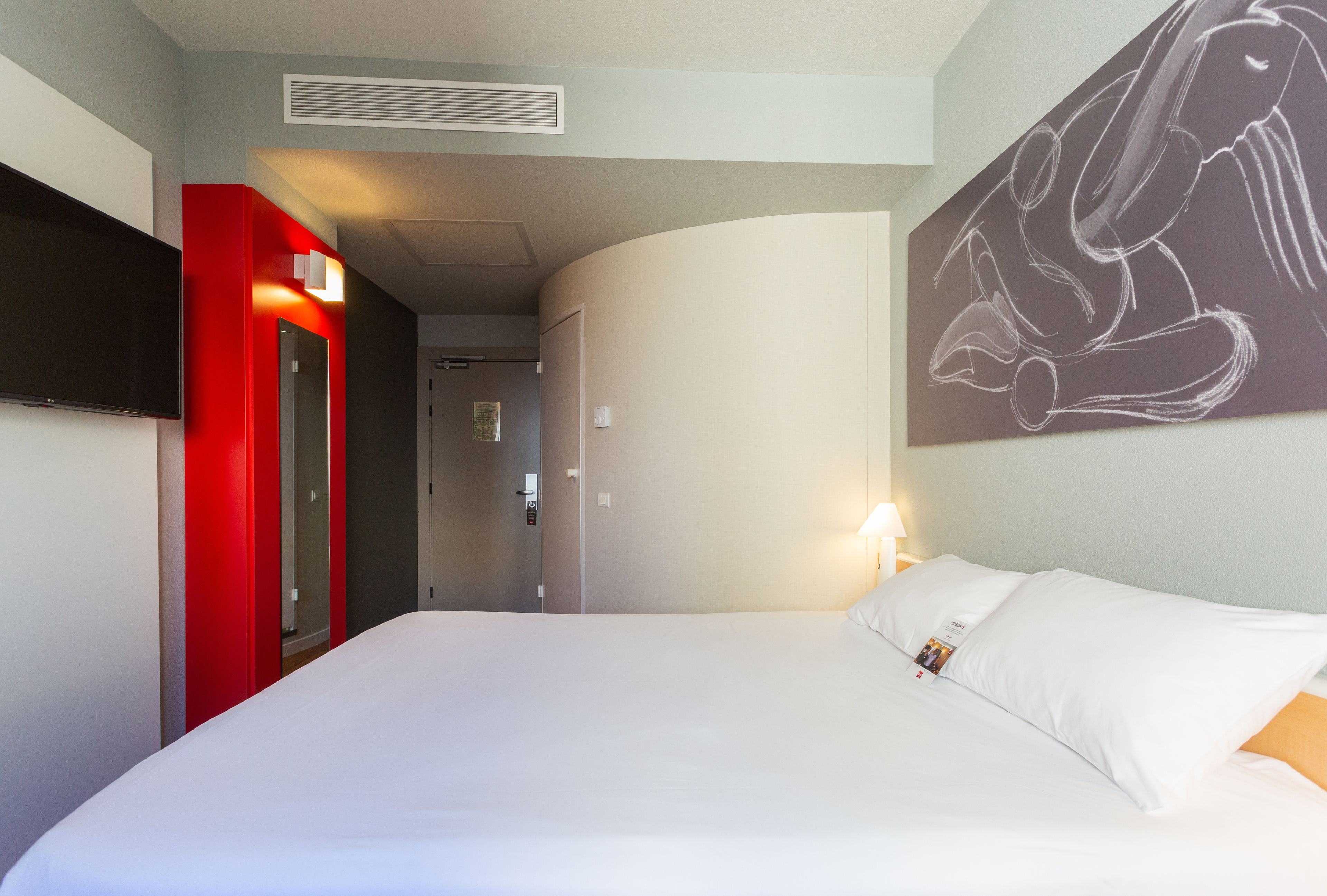 Una habitación de hotel con una cama, un televisor de pantalla plana montado en la pared, una puerta roja y un cuadro en la pared.