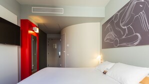 Ein Hotelzimmer mit Bett, einem flachbildfernseher an der Wand, einer roten Tür und einem Gemälde.