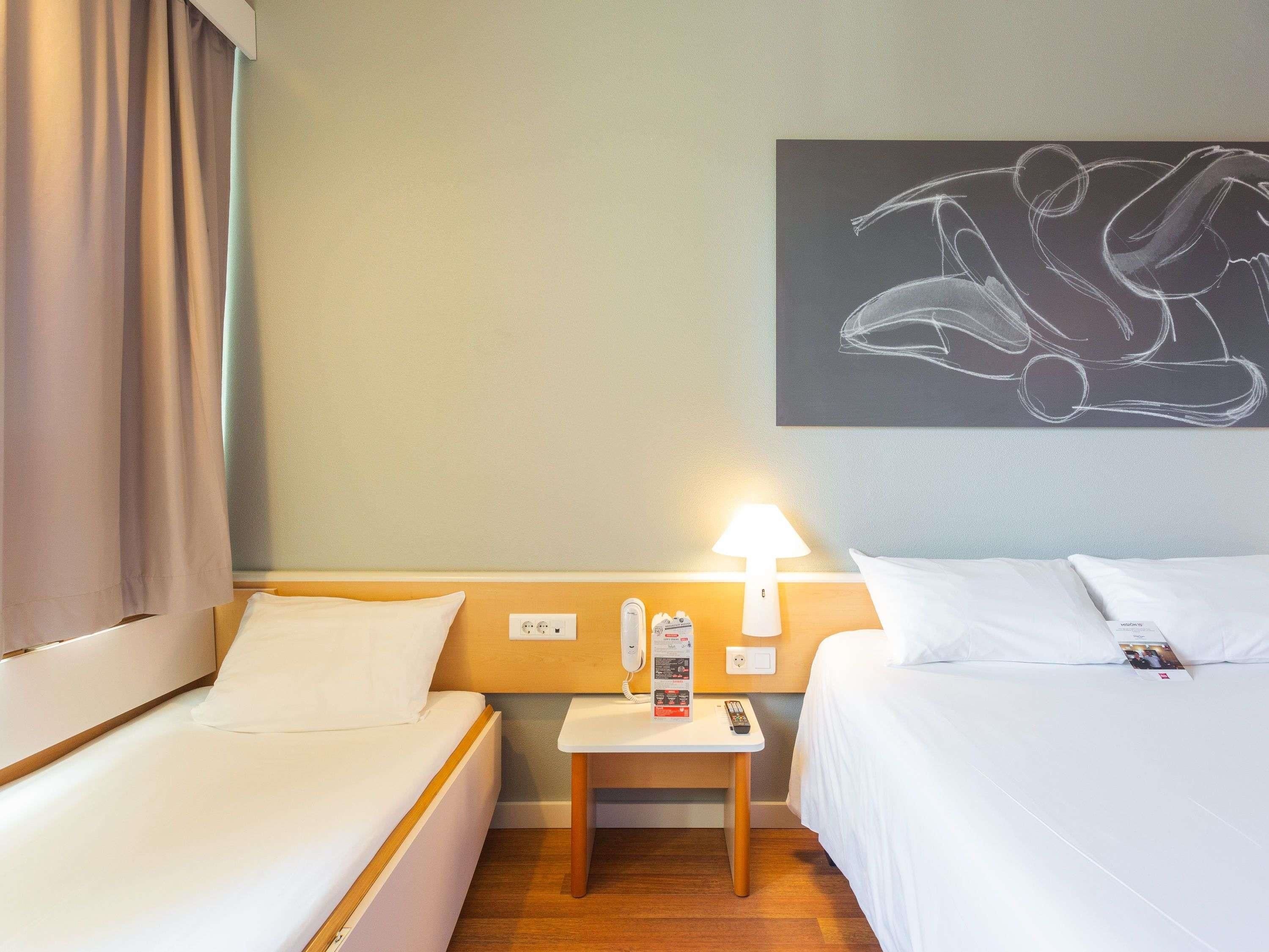 Una habitación de hotel con dos camas, una mesita de noche, una lámpara, un cuadro y una ventana con cortinas.