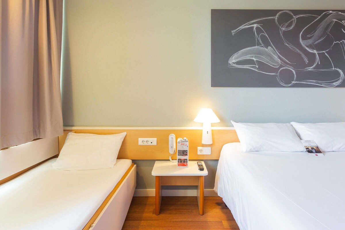 A hotel room with two beds, a nightstand, a lamp, a painting, and a window with curtains.