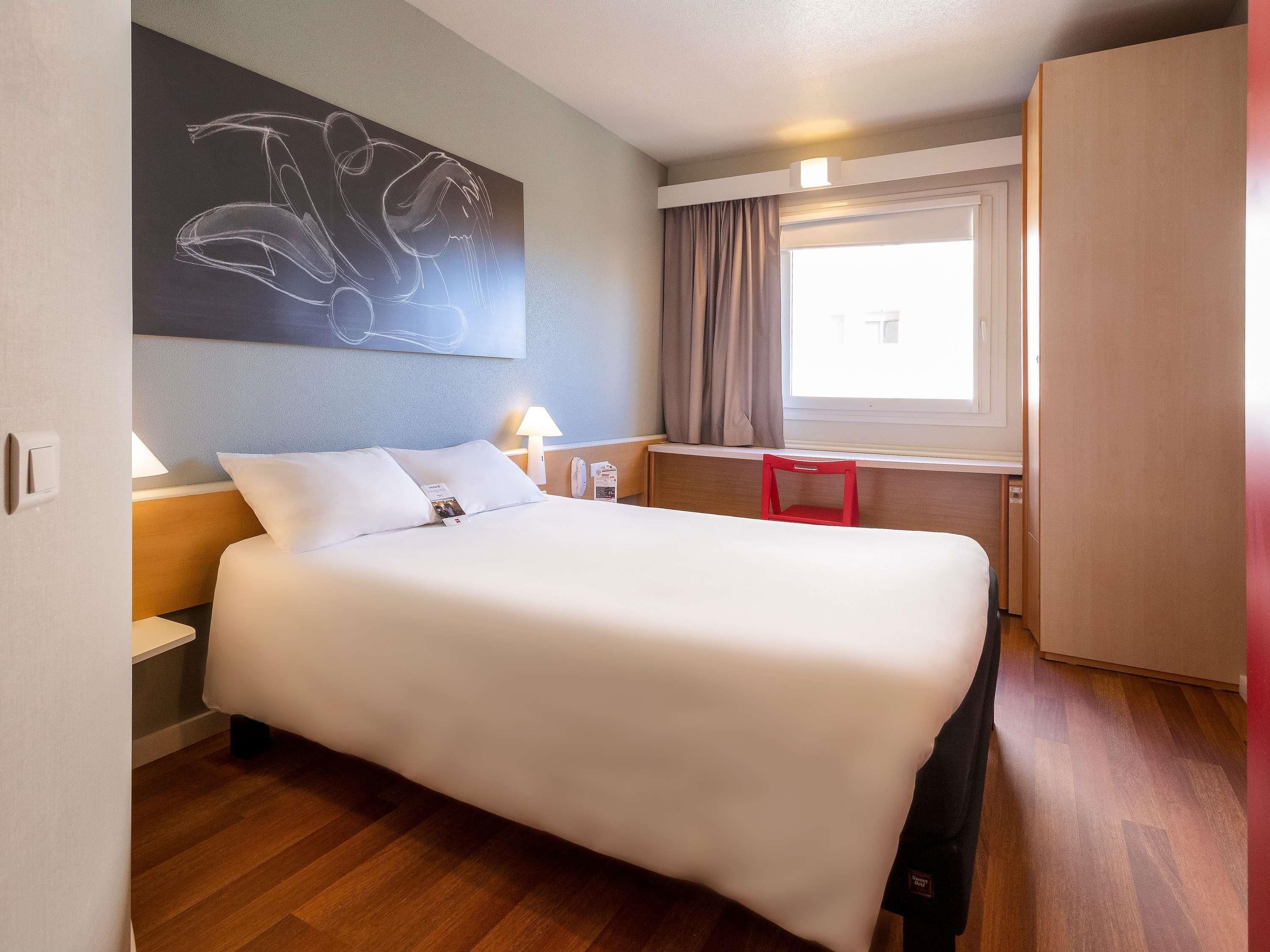 A hotel room with a bed, a desk, a window with curtains, and a wall art piece.