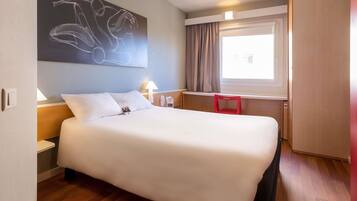 A hotel room with a bed, a desk, a window with curtains, and a wall art piece.
