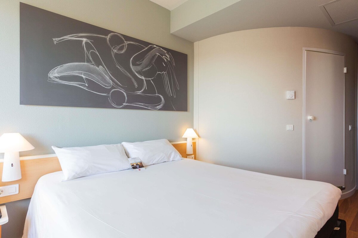 A hotel room with a bed, two bedside lamps, a wardrobe, and a large abstract painting on the wall.
