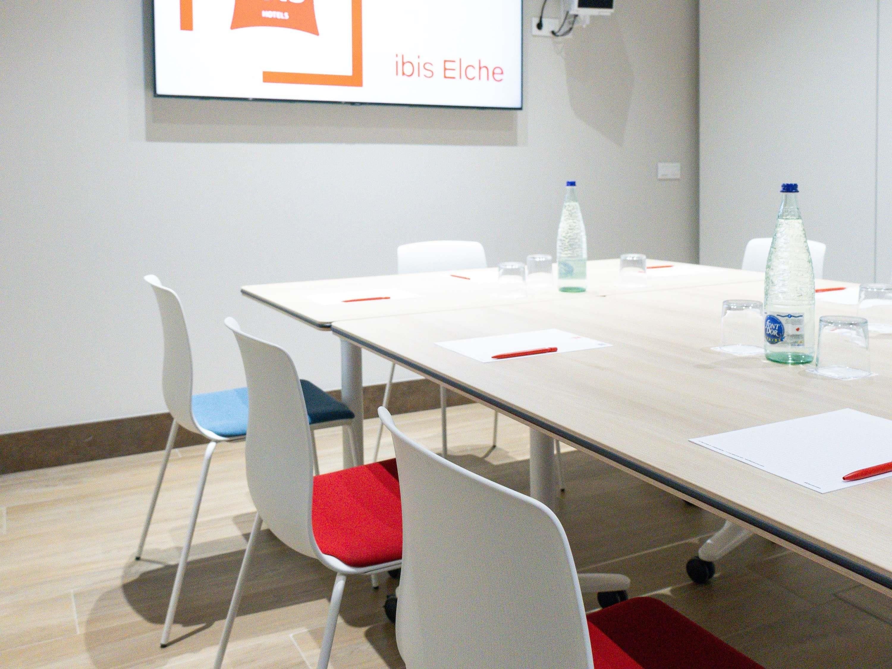 Uma sala de conferências com uma mesa comprida, cadeiras brancas com almofadas vermelhas e uma tela de projetor exibindo 'ibis Elche'.