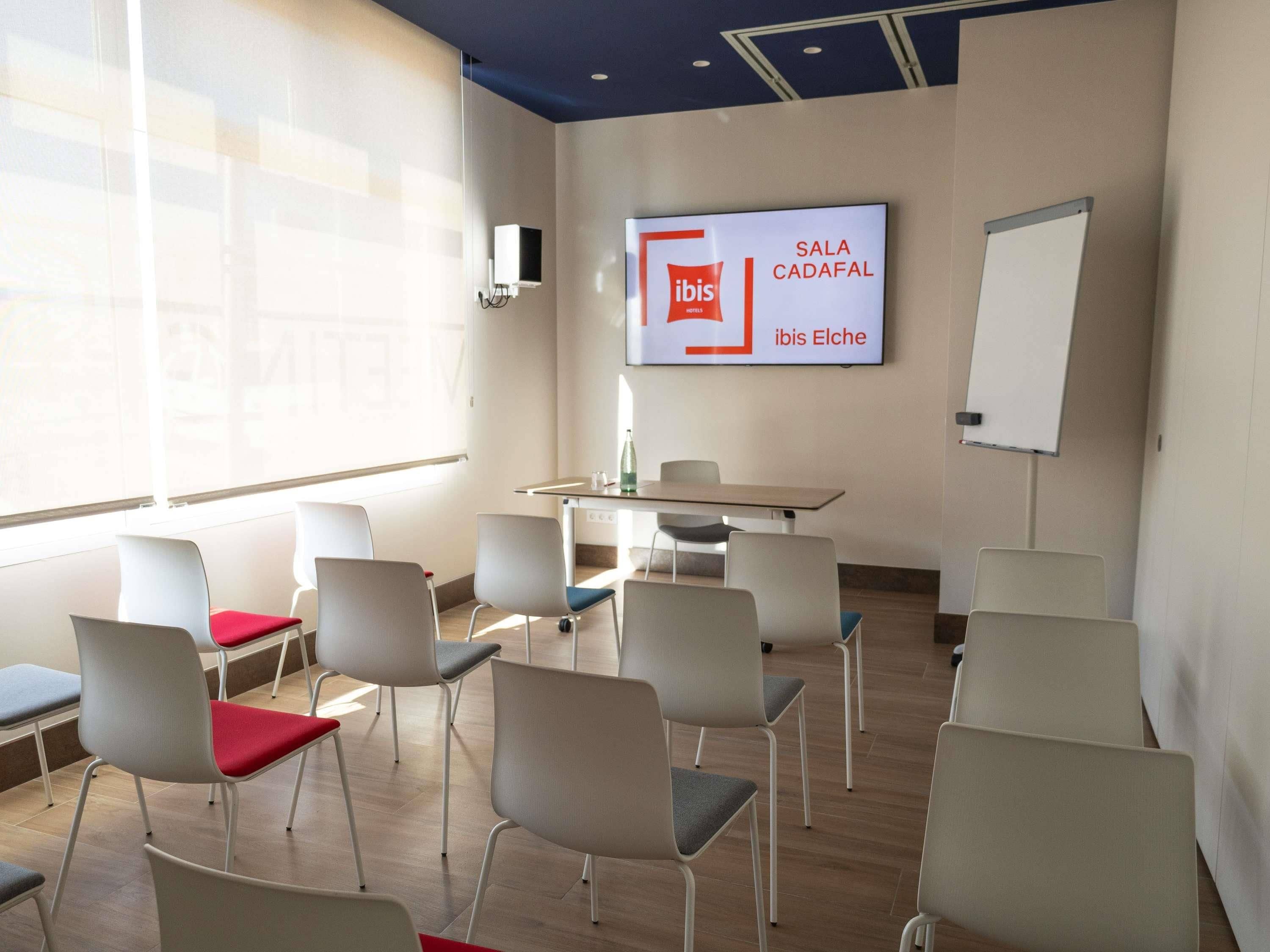Un salón de conferencias con sillas blancas, una mesa, una pizarra y una pantalla mostrando 'ibis HOTEL SALA CADAFAL ibis Elche'.